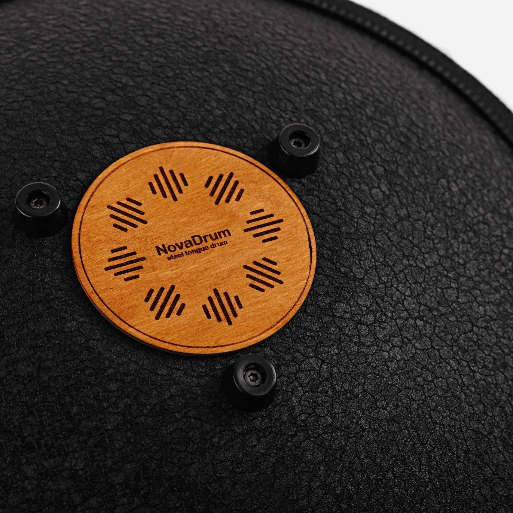 Close-up back view of Novadrum 38 steel tongue drum in black textured finish, focusing on the wooden cover with engraved ‘NovaDrum’ branding, rubber feet, and black rope rim.