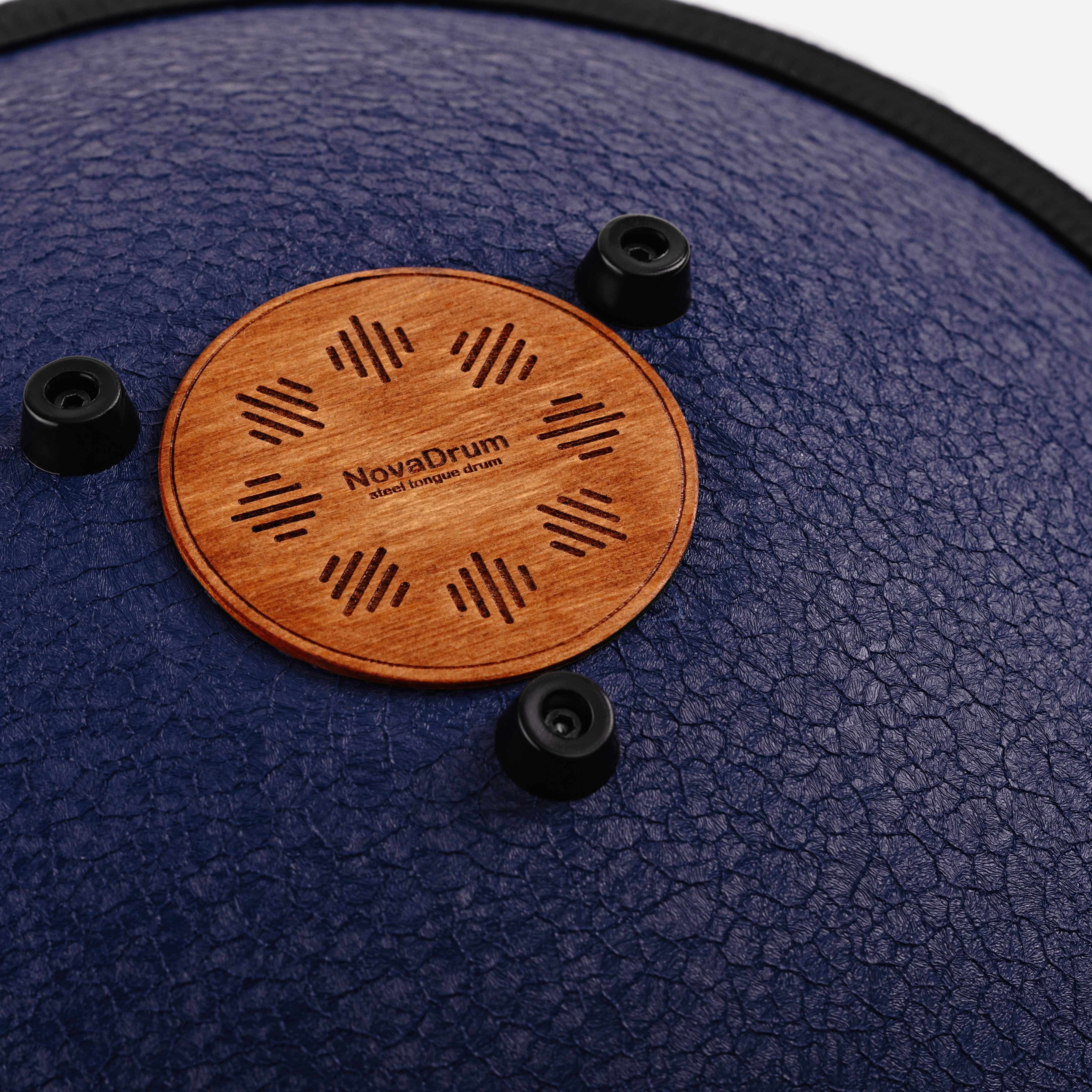 Close-up back view of Novadrum 38 steel tongue drum in blue textured finish, focusing on the wooden cover with engraved ‘NovaDrum’ branding, rubber feet, and black rope rim.